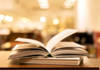 Open book on a table in a warmly lit library, pages fanned out. The blurred background suggests rows of bookshelves, evoking a cozy, inviting atmosphere.